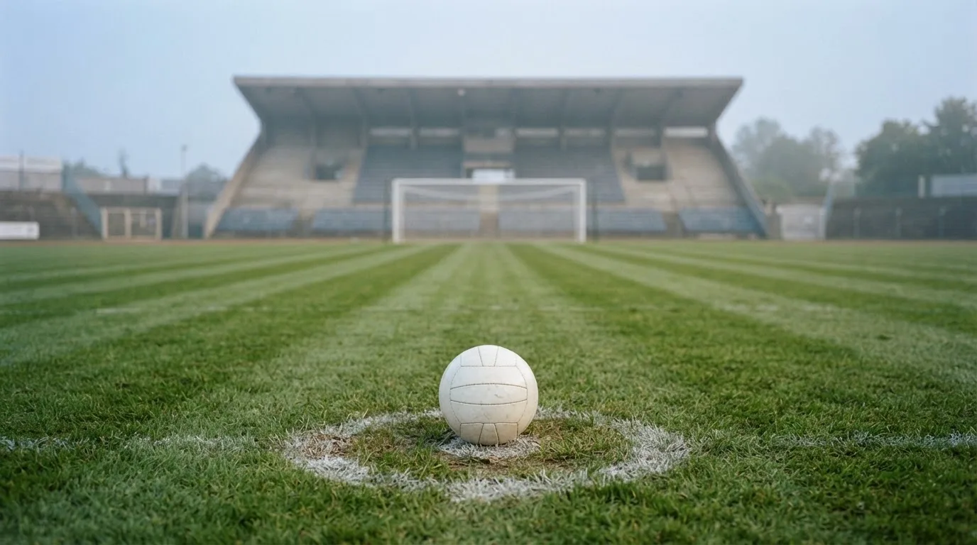 Ein Fußball liegt auf dem Elfmeterpunkt eines leeren Rasenplatzes, im Hintergrund eine Tribüne im Dunst.