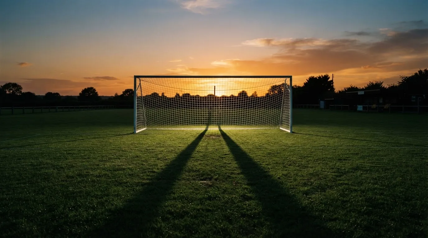 Ein Fußballtor mit Netz bei Sonnenuntergang auf einem leeren Platz, die Schatten der Torpfosten liegen lang auf dem Gras.