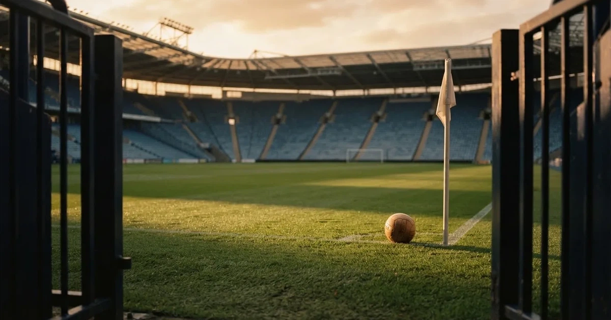 Blick durch ein Stadiontor auf einen grünen Rasen im Abendlicht, ein Ball ruht an der Eckfahne, die Tribünen sind leer.