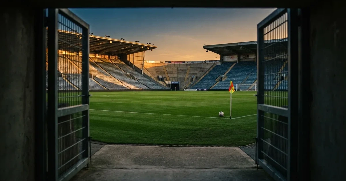 Blick durch ein Stadiontor auf einen grünen Rasen im Abendlicht, ein Ball liegt an der Eckfahne, die Ränge sind leer.