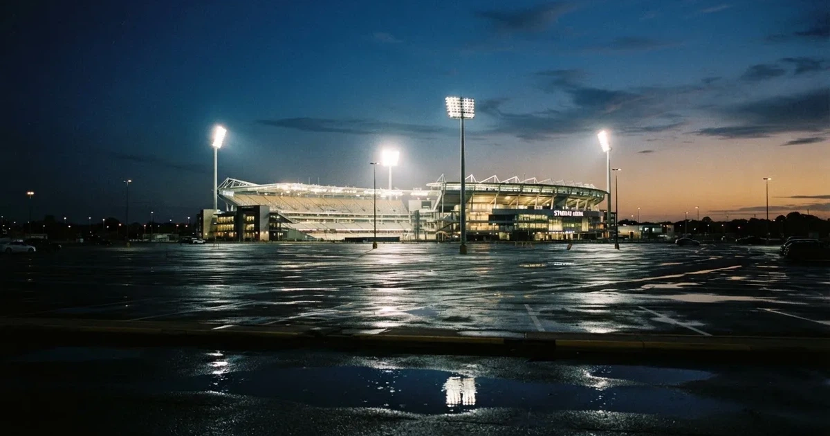 Ein Stadion unter Flutlicht aus großer Entfernung gesehen, davor ein regennasser Parkplatz in der Dämmerung.