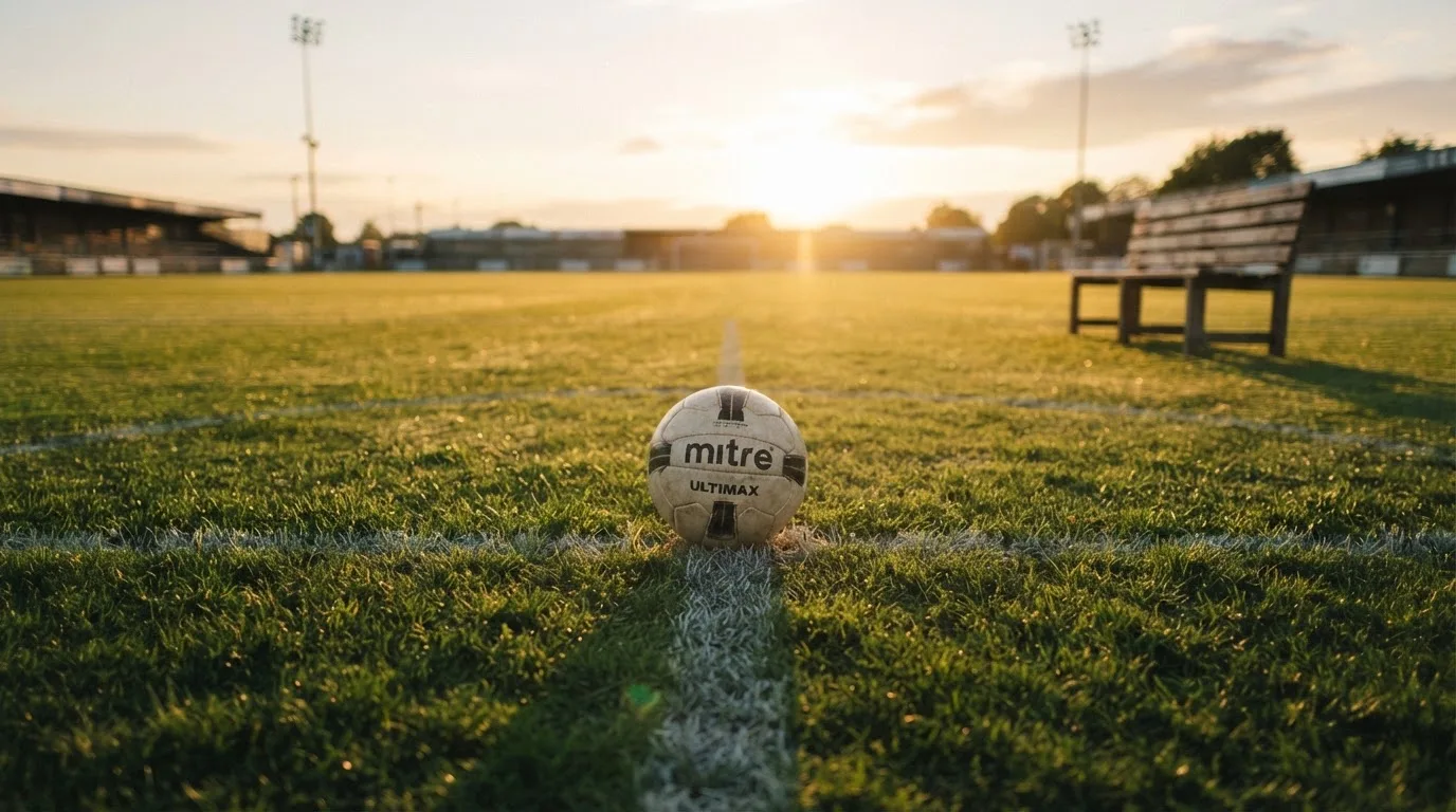 Ein Fußball auf einem Rasenplatz im Gegenlicht der tiefstehenden Sonne, am Spielfeldrand eine leere Ersatzbank.