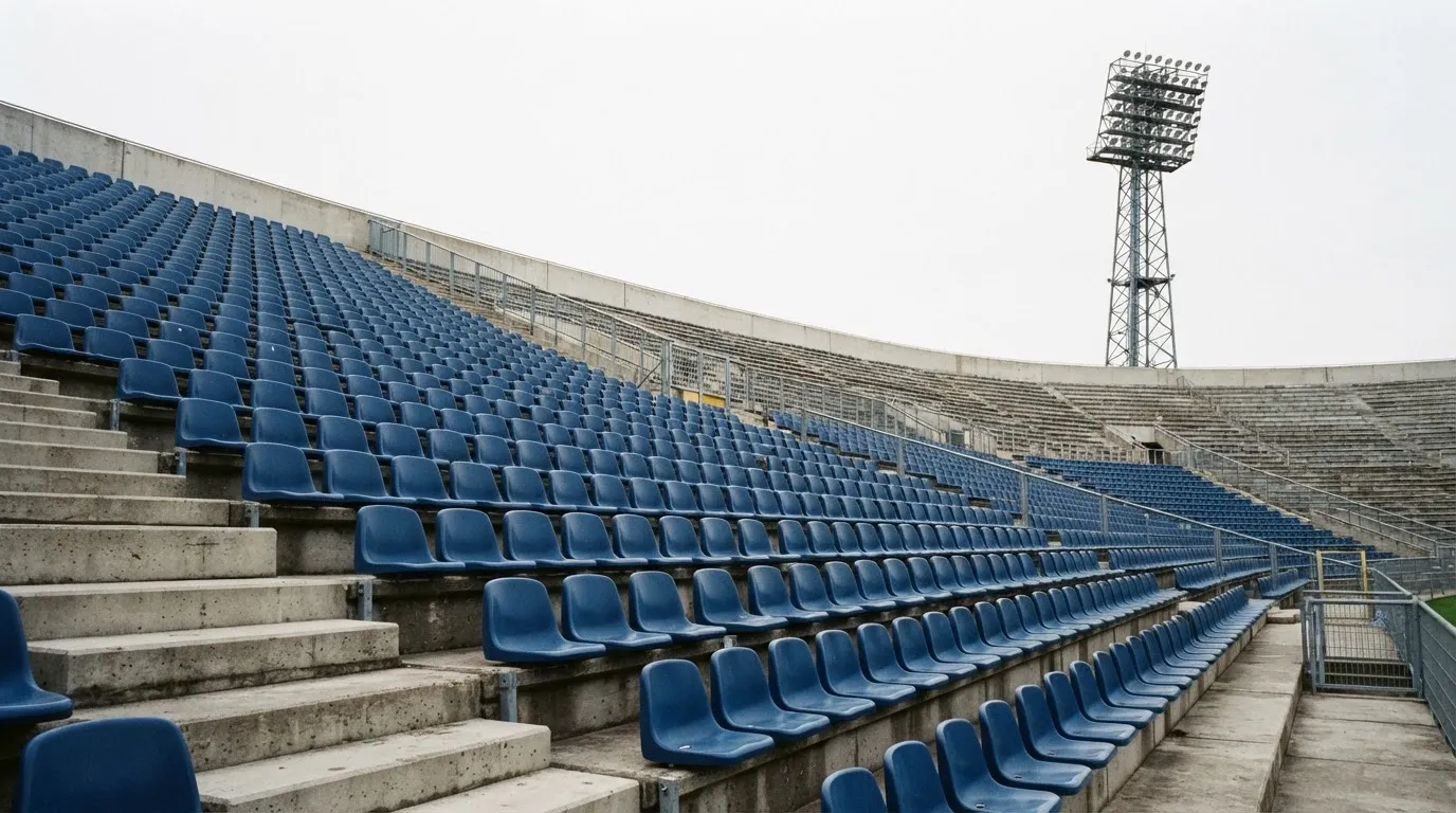 Eine leere Tribüne eines Fußballstadions bei Tageslicht, Sitzschalen in Reihen und dahinter ein Flutlichtmast.