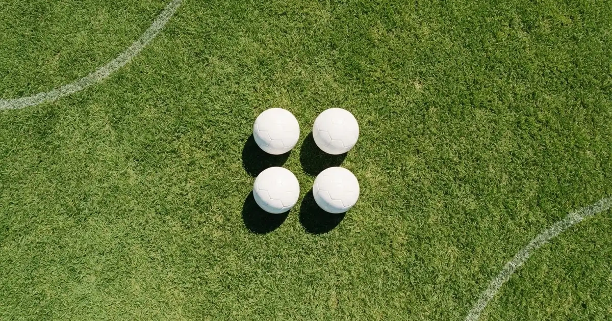 Vier Fußbälle in einem Quadrat auf einem Rasenplatz, von oben fotografiert, mit harten Mittagsschatten.