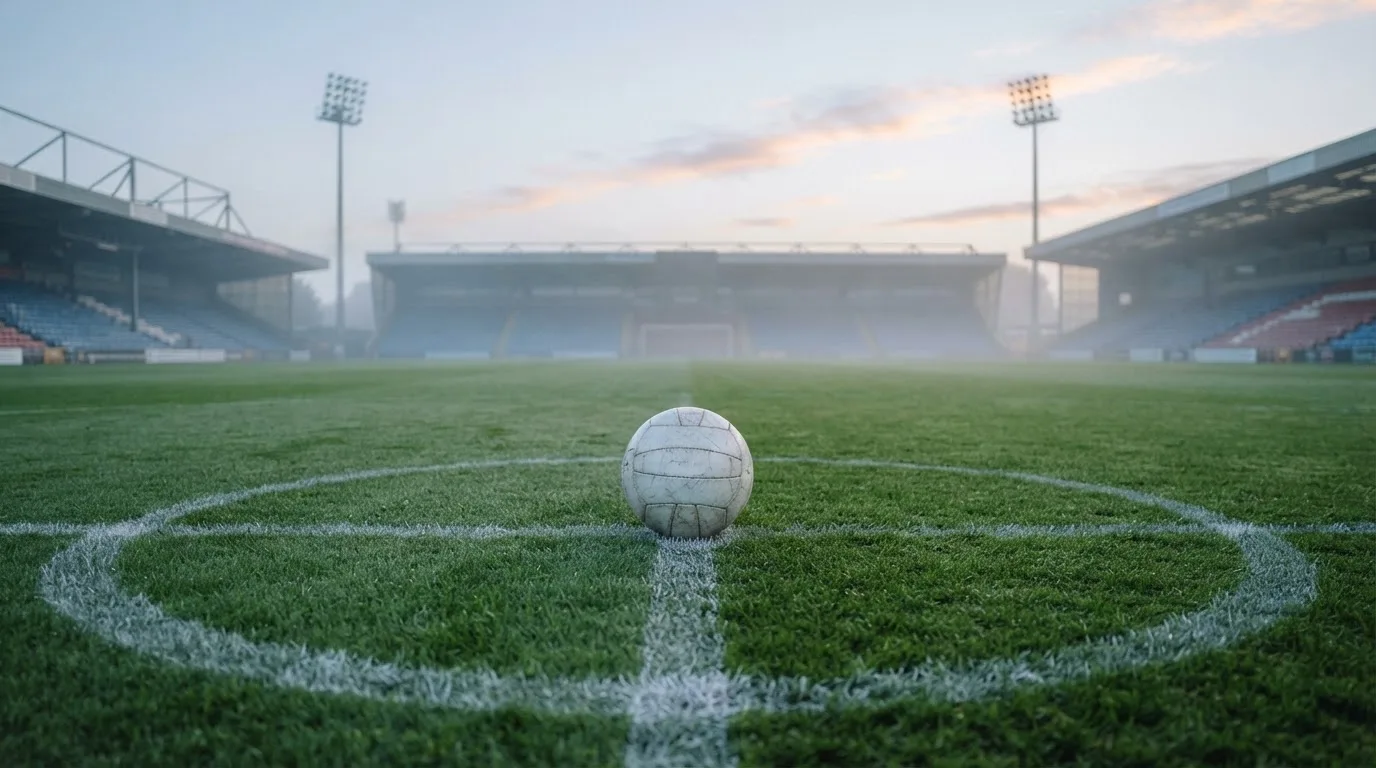 Ein Fußball liegt auf dem Anstoßpunkt eines leeren Rasenplatzes, im Hintergrund hängt Morgendunst über den Tribünen.