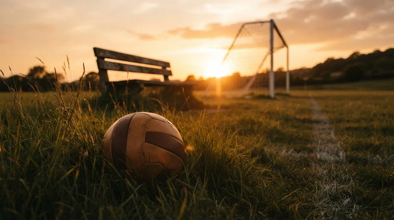 Ein Fußball liegt im Gras am Spielfeldrand, dahinter eine leere Bank und ein Tor im Gegenlicht.