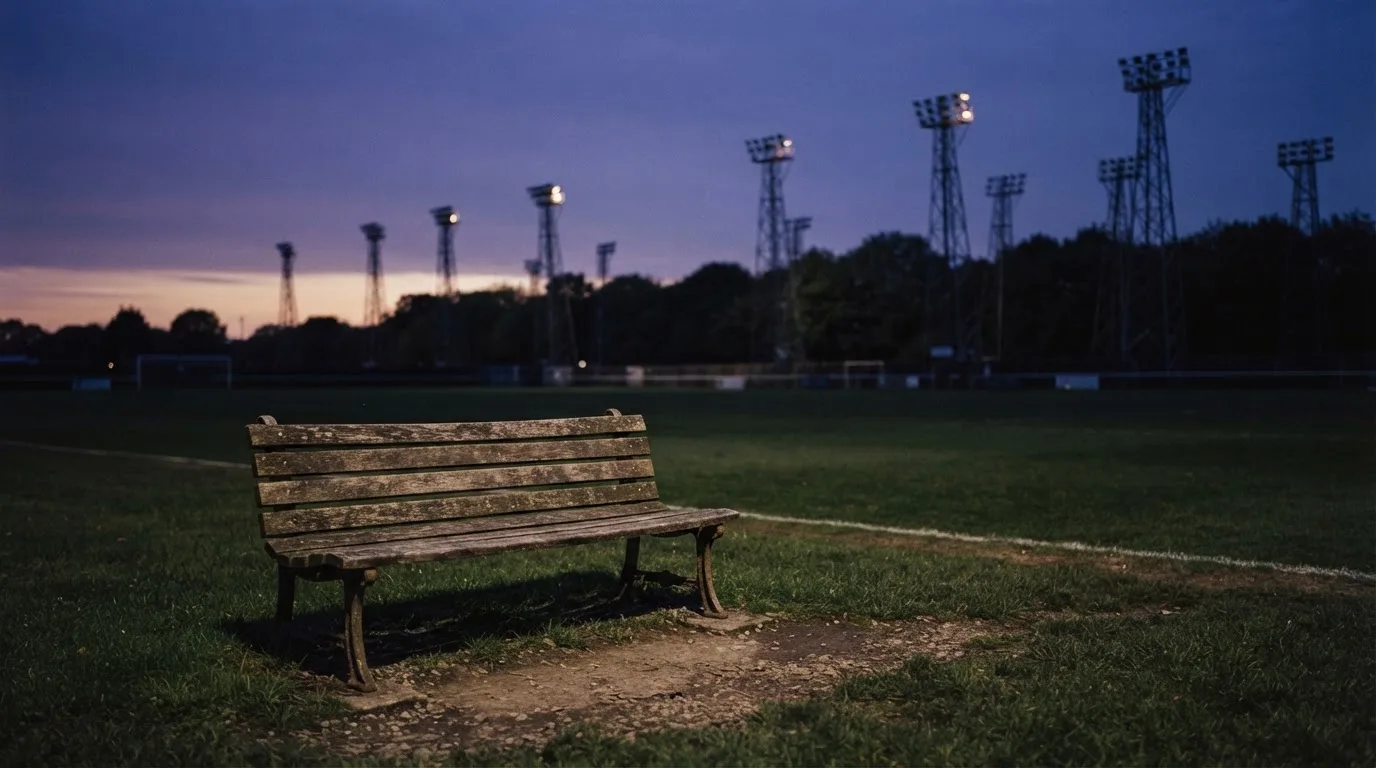 Eine leere Parkbank am Rand eines Fußballfelds in der Abenddämmerung, im Hintergrund ragen dunkle Flutlichtmasten in den Himmel.