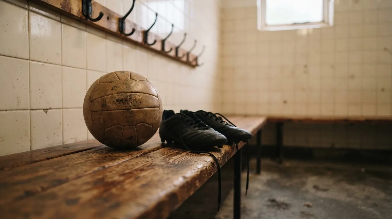 Ein Fußball liegt neben einem Paar Fußballschuhen ohne Markenzeichen auf einer Holzbank in einer leeren Umkleide.