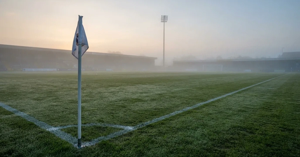Eine Eckfahne steht im Nebel am Rand eines leeren Rasenplatzes in der Morgendämmerung, dahinter ein Flutlichtmast.