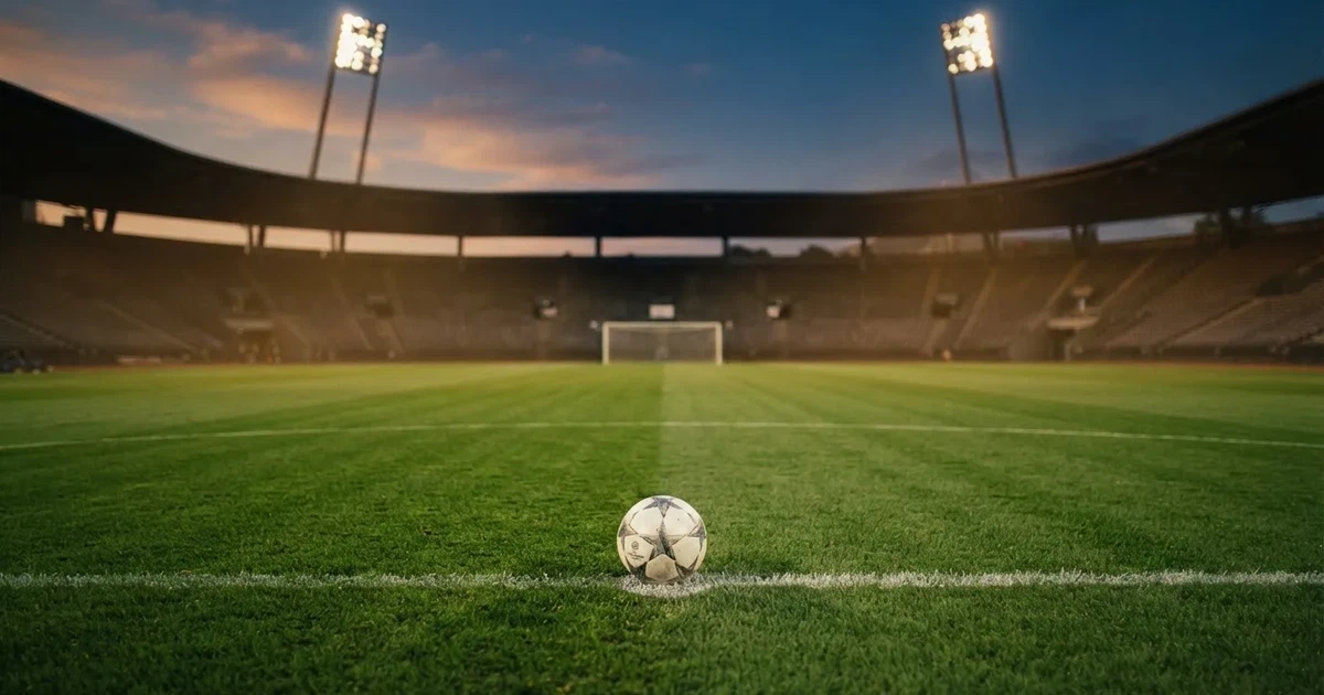 Ein Fußballplatz mit frisch gemähtem Rasen im Abendlicht, ein Ball liegt im Strafraum, im Hintergrund unscharf eine leere Tribüne.