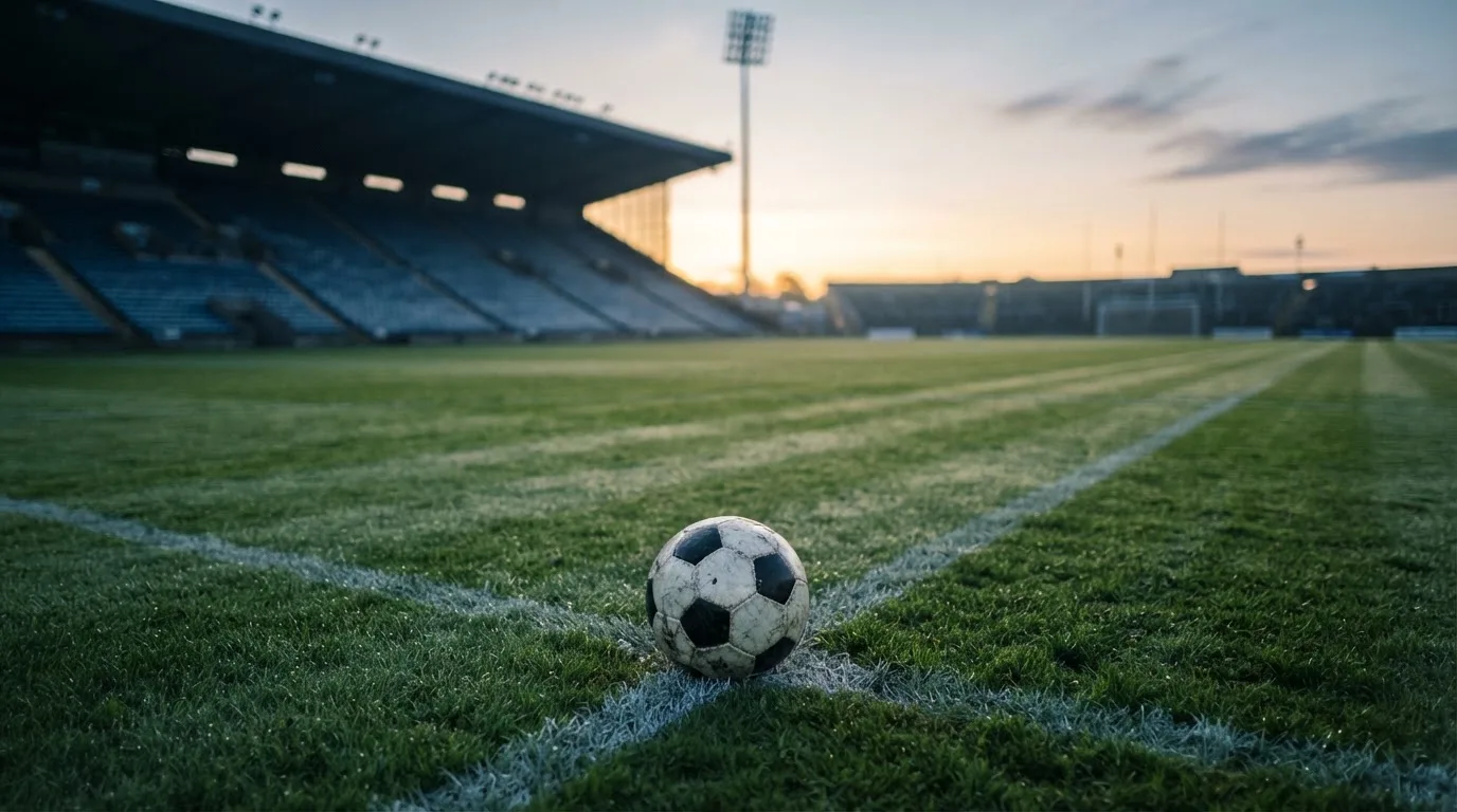 Ein Fußball liegt auf dem Anstoßpunkt eines leeren Rasenplatzes im frühen Morgenlicht, dahinter eine leere Tribüne.