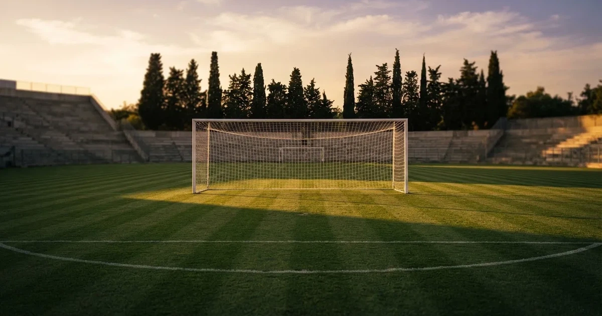 Ein Fußballtor mit Netz auf einem leeren Platz im Abendlicht, der Rasen frisch gemäht, im Hintergrund Zypressen.