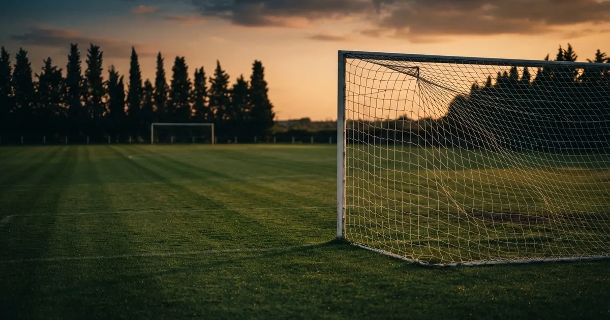 Ein Fußballtor mit Netz auf einem leeren Platz im Abendlicht, der Rasen frisch gemäht, im Hintergrund Zypressen.