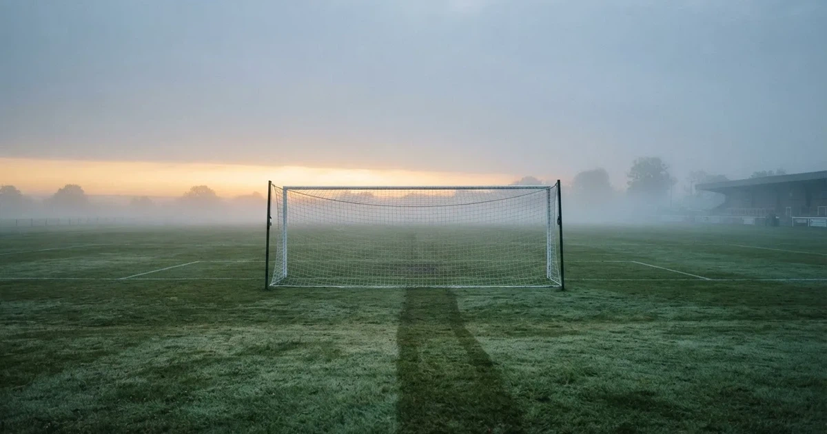 Ein Fußballtor mit Netz steht auf einem leeren Platz im Nebel der Morgendämmerung, der Rasen ist taufeucht.