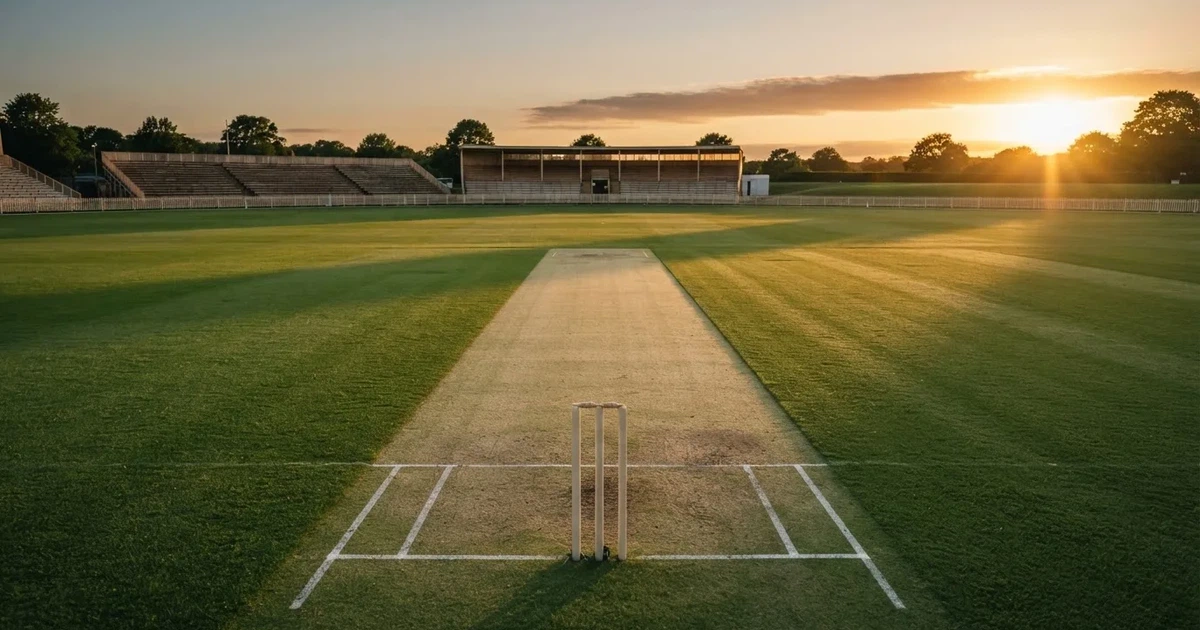 Ein weites grünes Cricketfeld mit einem hellen Streifen in der Mitte, tief stehende Sonne, im Hintergrund eine leere Tribüne.