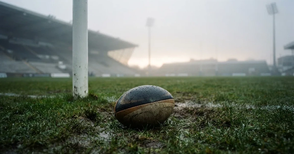 Ein ovaler Rugbyball liegt vor den hohen Torstangen auf nassem Rasen, über dem Stadion zieht Nebel auf.