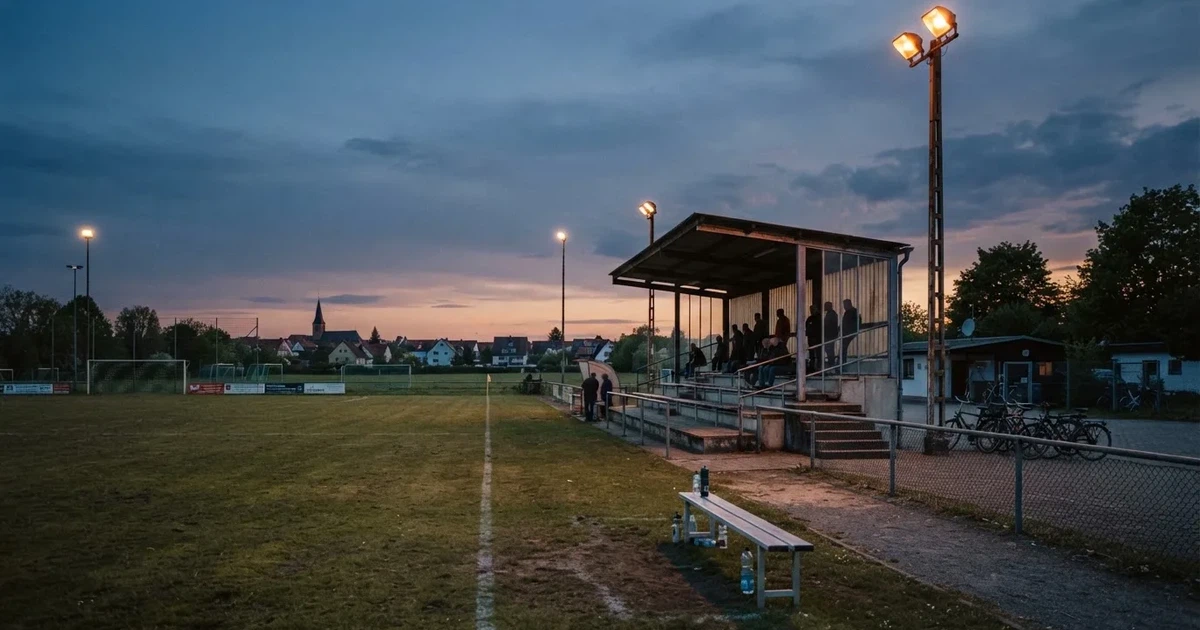 Ein einfacher Rasenplatz mit kleiner Stehtribüne und Flutlichtmasten in der Dämmerung, auf der Tribüne sitzen nur wenige Zuschauer.