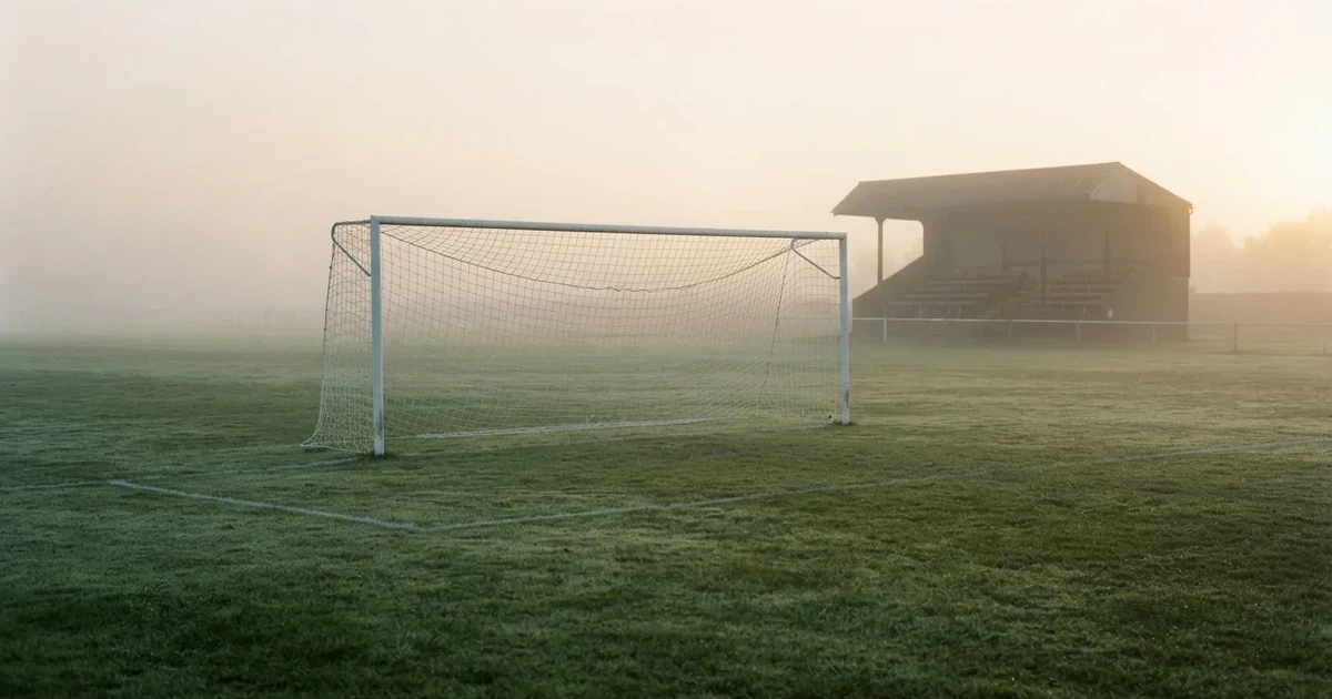 Ein Fußballtor mit Netz steht im Morgennebel auf einem leeren Rasenplatz, im Hintergrund die Silhouette einer kleinen Tribüne.