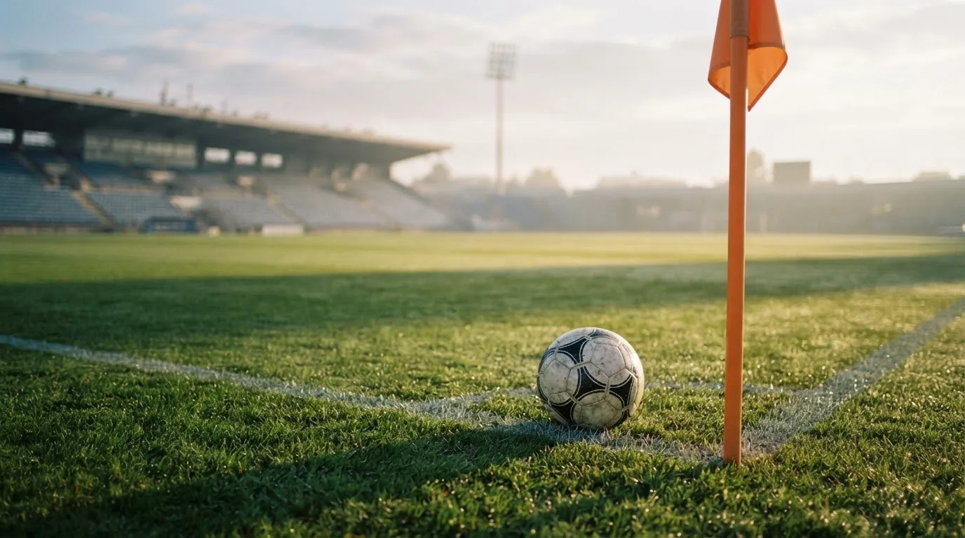 Ein Fußball liegt auf Rasen neben einer Eckfahne ohne Aufdruck auf einem leeren Platz im Morgenlicht.