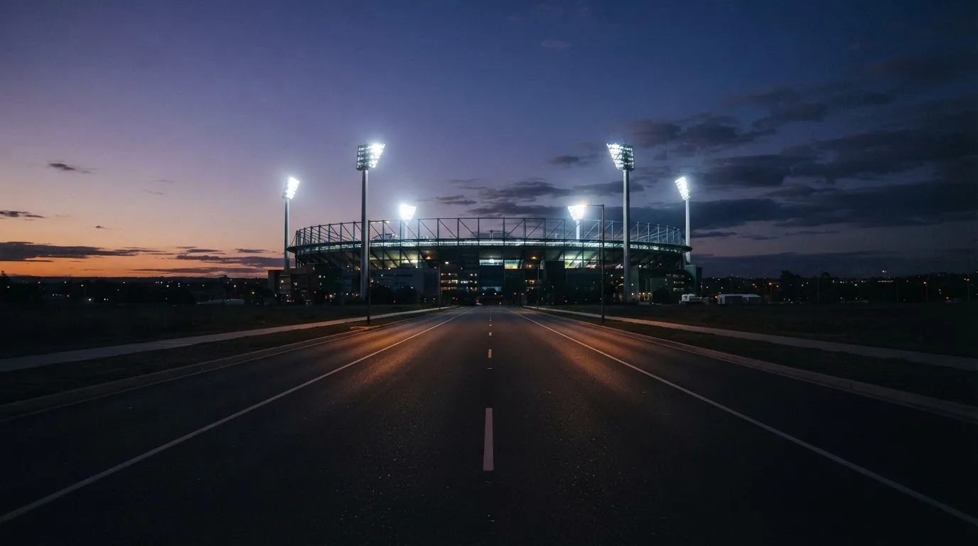 Ein Stadion unter Flutlicht aus großer Entfernung, davor eine leere Straße in der Abenddämmerung.