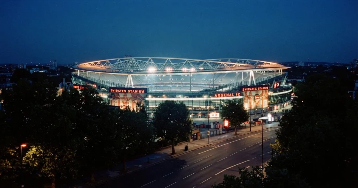Ein Fußballstadion bei Nacht aus großer Entfernung, hell erleuchtet unter Flutlicht, davor eine dunkle Straße und Bäume.