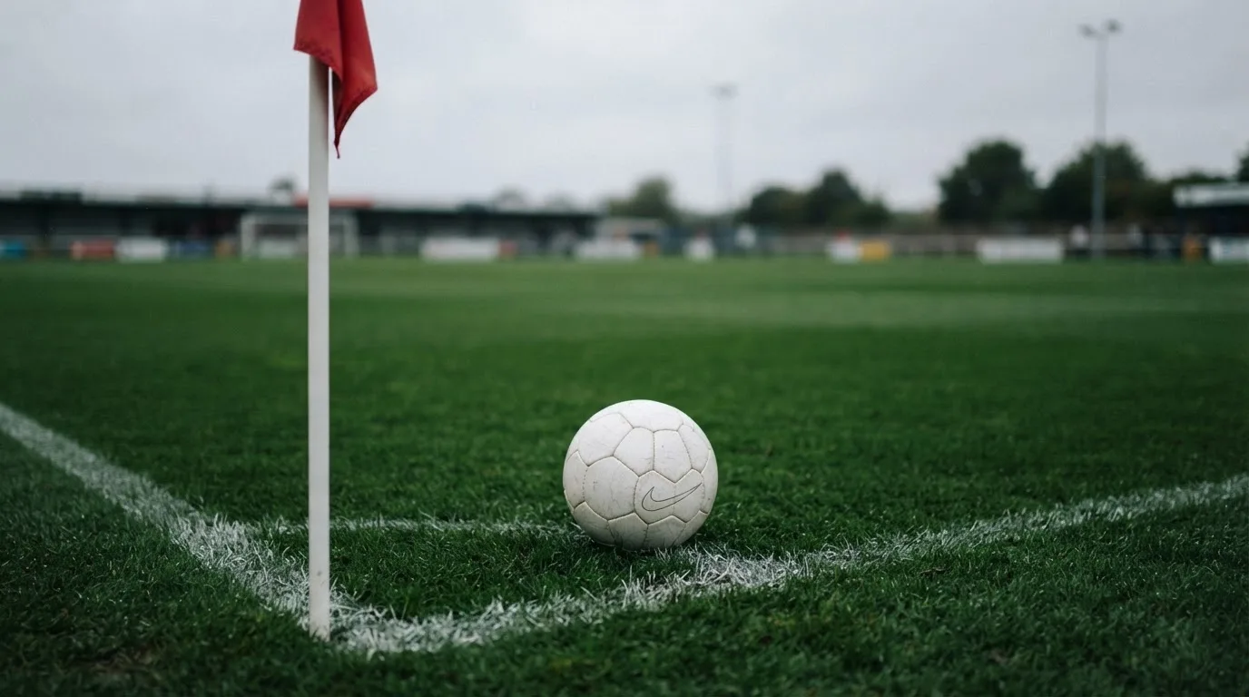 Ein Fußball liegt neben einer Eckfahne auf kurz gemähtem Rasen unter bewölktem Himmel.